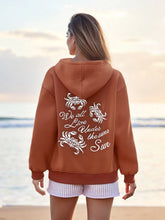 Person wearing a brown hoodie with a quote on the back, standing on a beach.